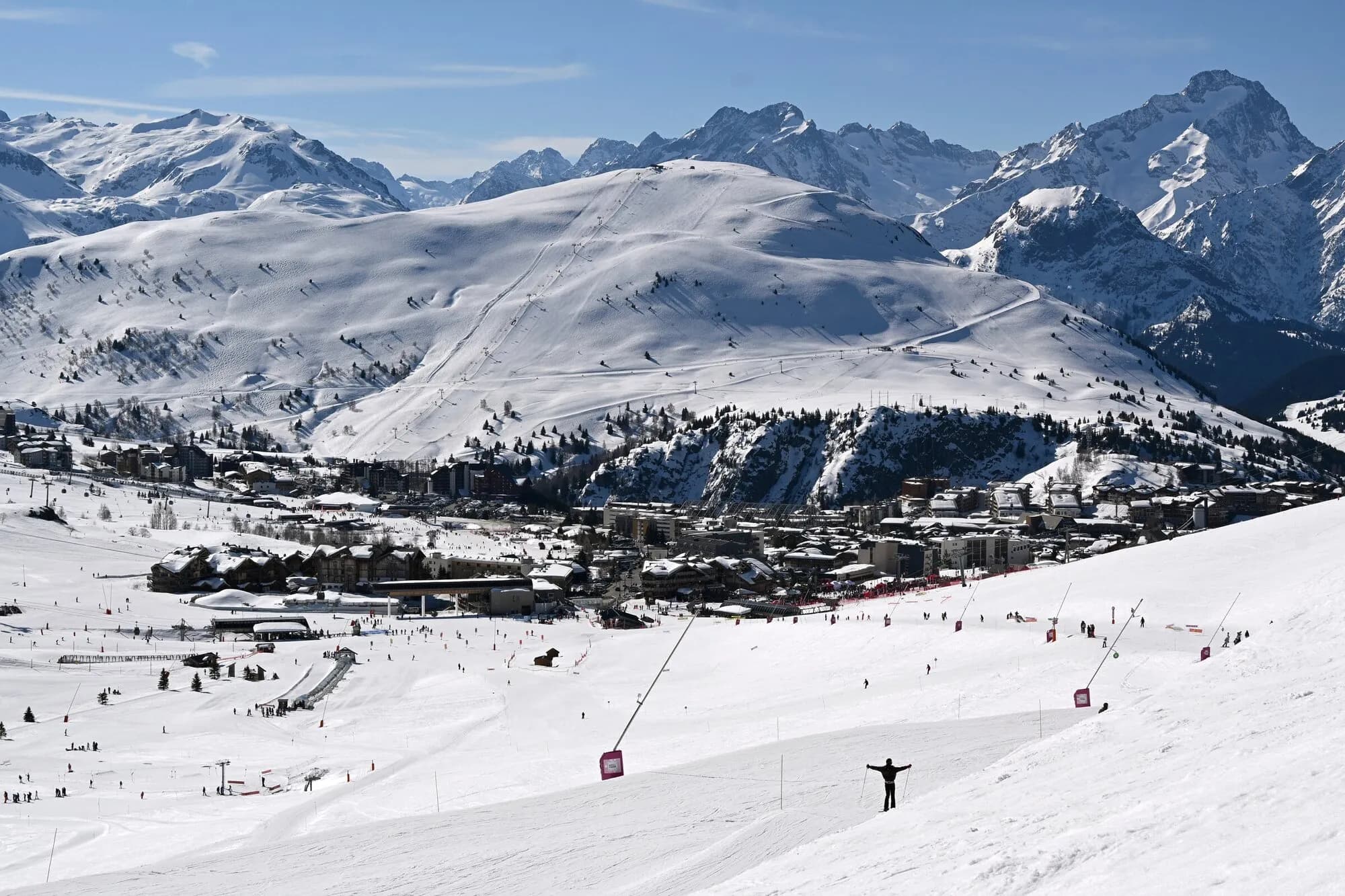 The resort spread across a snowy plateau below ski slopes and jagged peaks, with a skier in front.