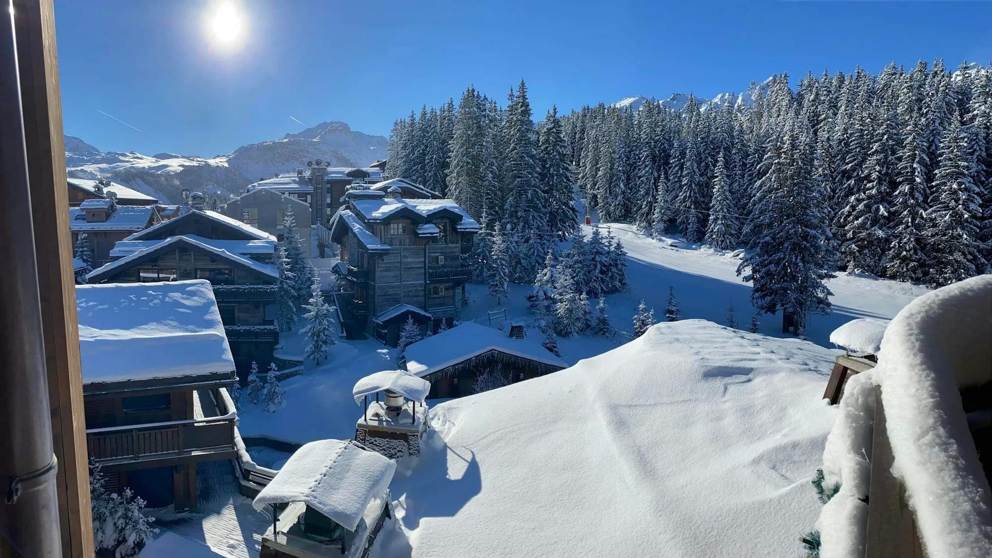 View from a window over deep-snow chalet roofs and a fir forest under bright sun.