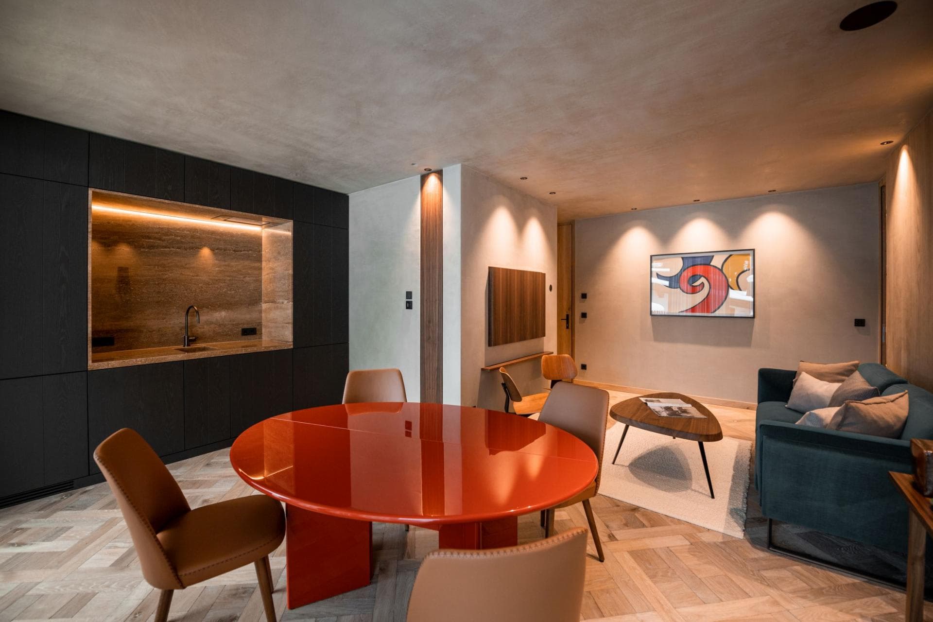 Open-plan living area with round red dining table seating 4, lounge with teal sectional, and wet bar with backlit shelving