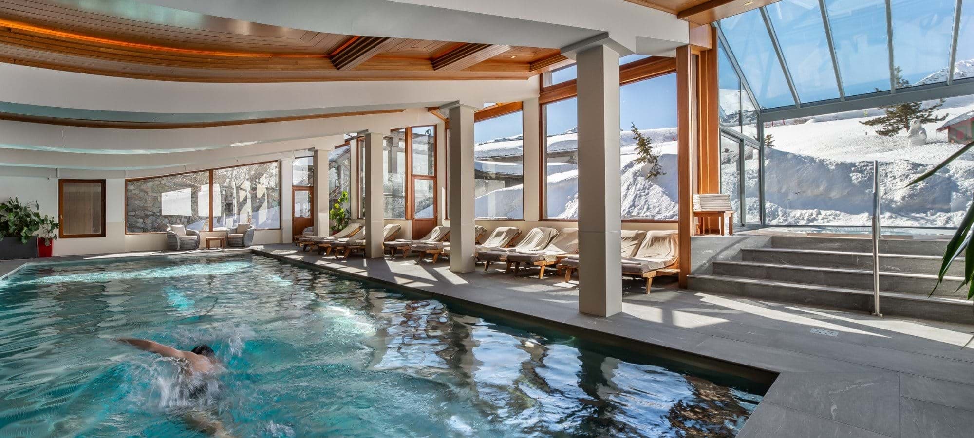 Indoor heated pool with poolside loungers and floor-to-ceiling mountain views