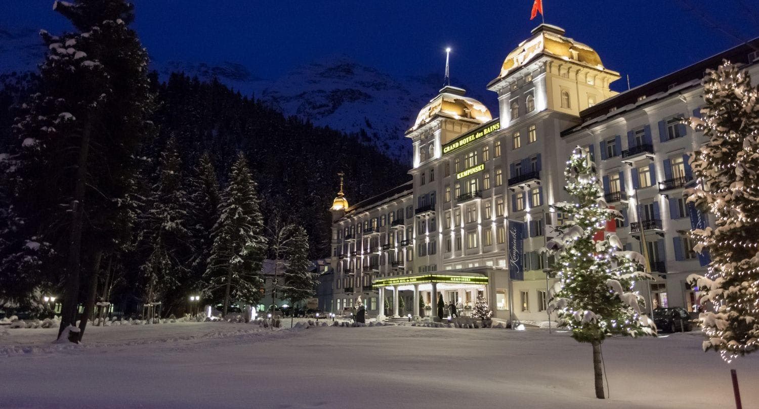 Hotel exterior at dusk with snow-covered grounds and mountain backdrop