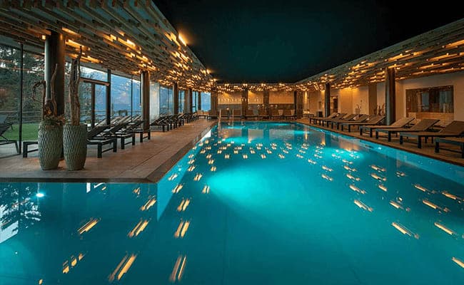 Indoor heated pool with mountain-facing floor-to-ceiling windows and poolside loungers