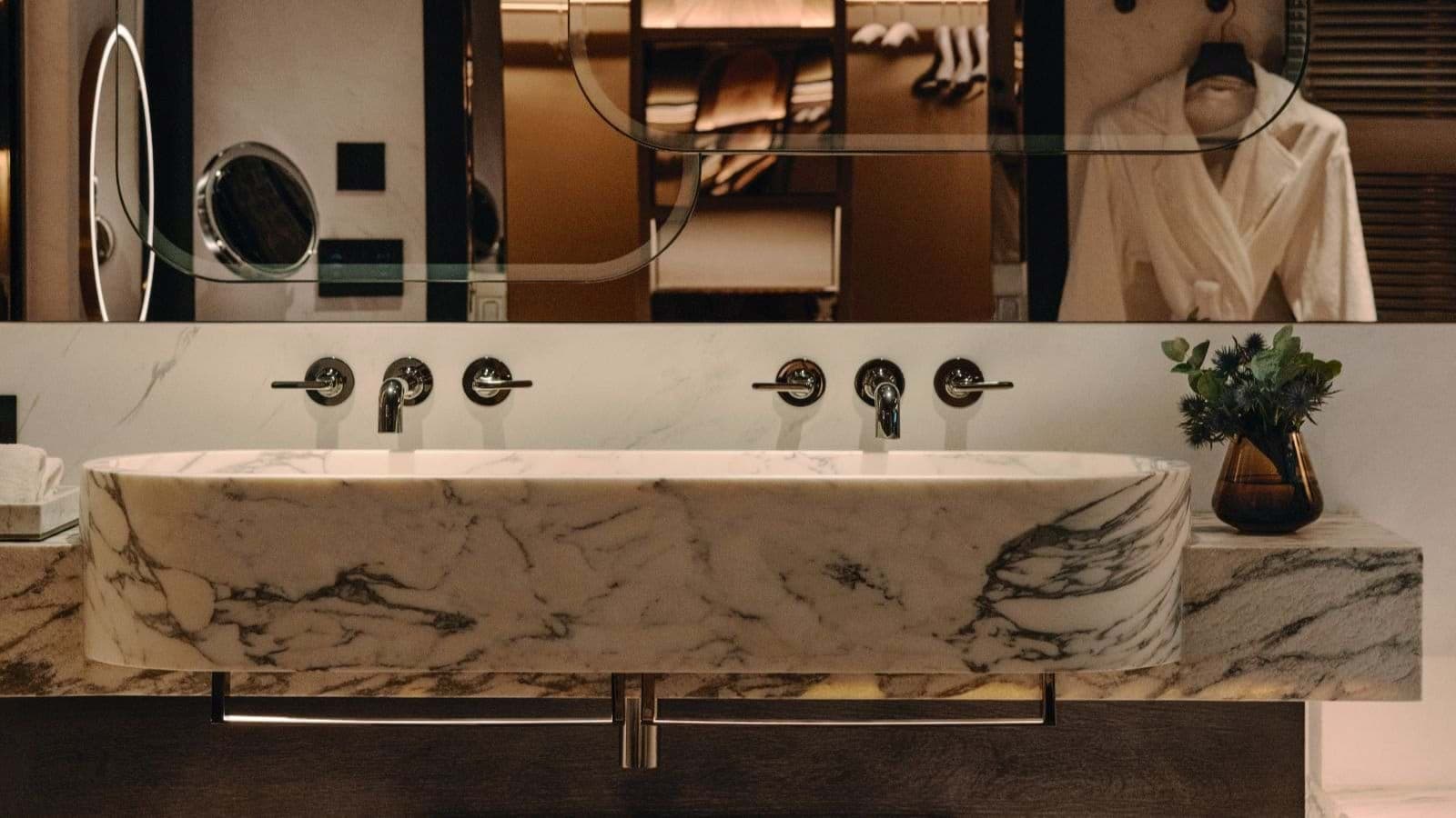 Bathroom vanity with marble trough sink, dual faucets, and walk-in closet beyond