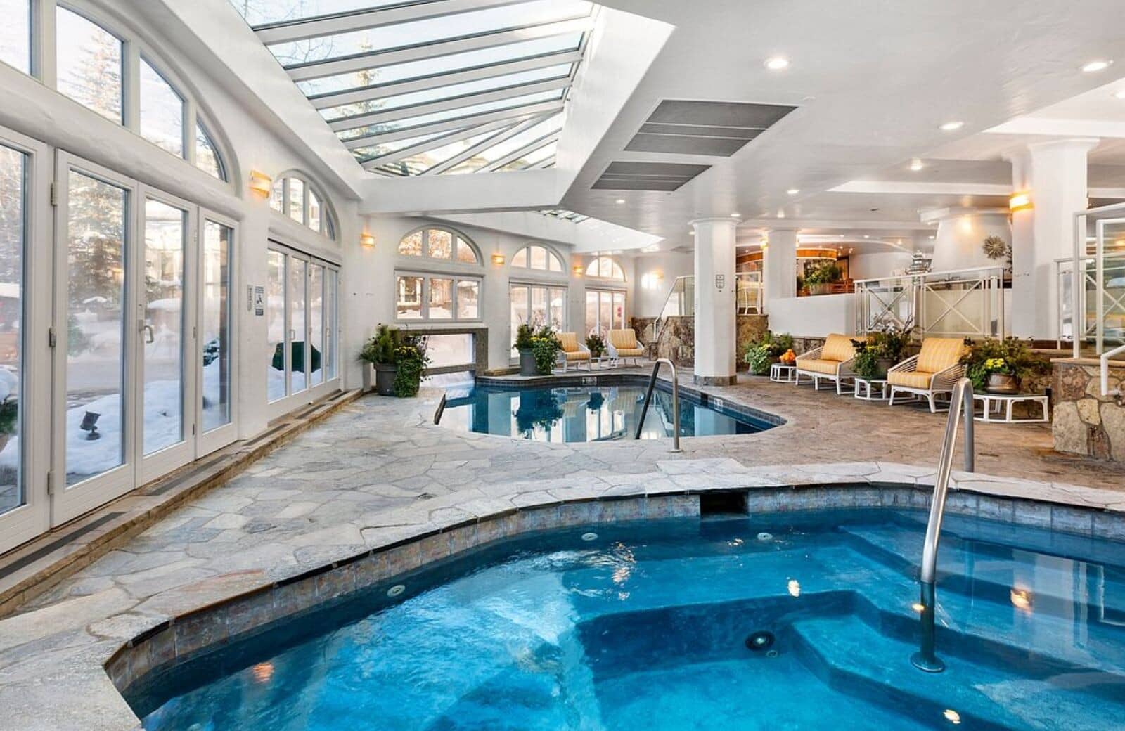 Indoor pool and hot tub with glass atrium ceiling and lounge seating