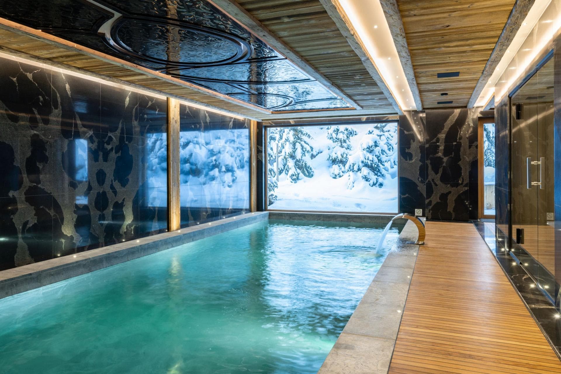 Indoor swimming pool with waterfall feature and snow-covered forest views