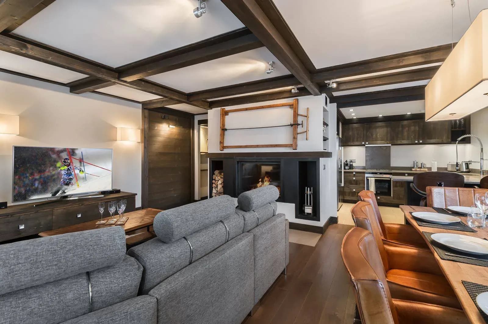 Living room with grey sofa, wall-mounted TV and wood fireplace, open to kitchen