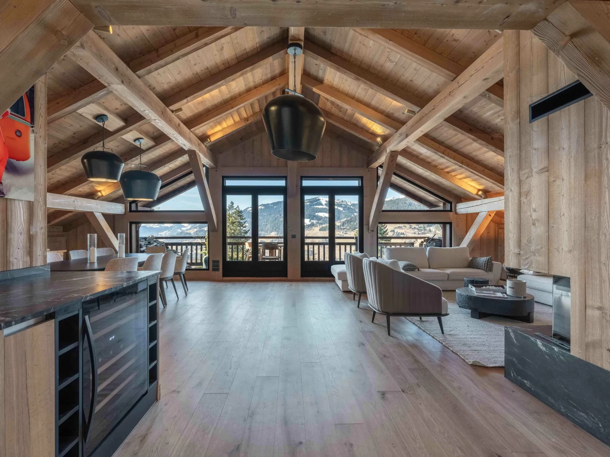 Open-plan living area under exposed beams with lounge, dining table and balcony doors to mountain views