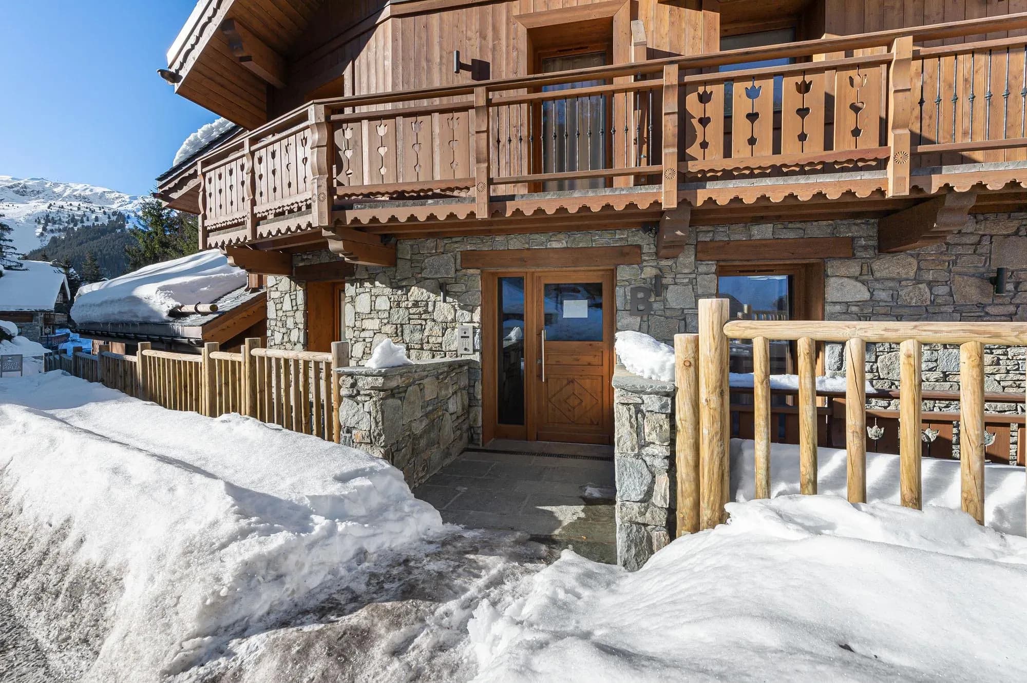 Chalet entrance with wooden door, stone walls and balcony above, snow-covered approach