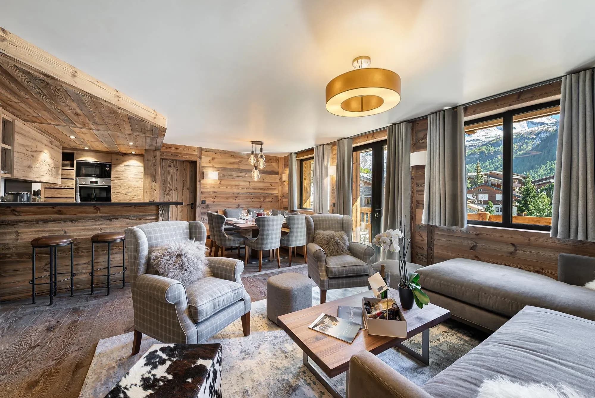 Open-plan living area with grey sofas, plaid armchairs and mountain views, kitchen bar behind