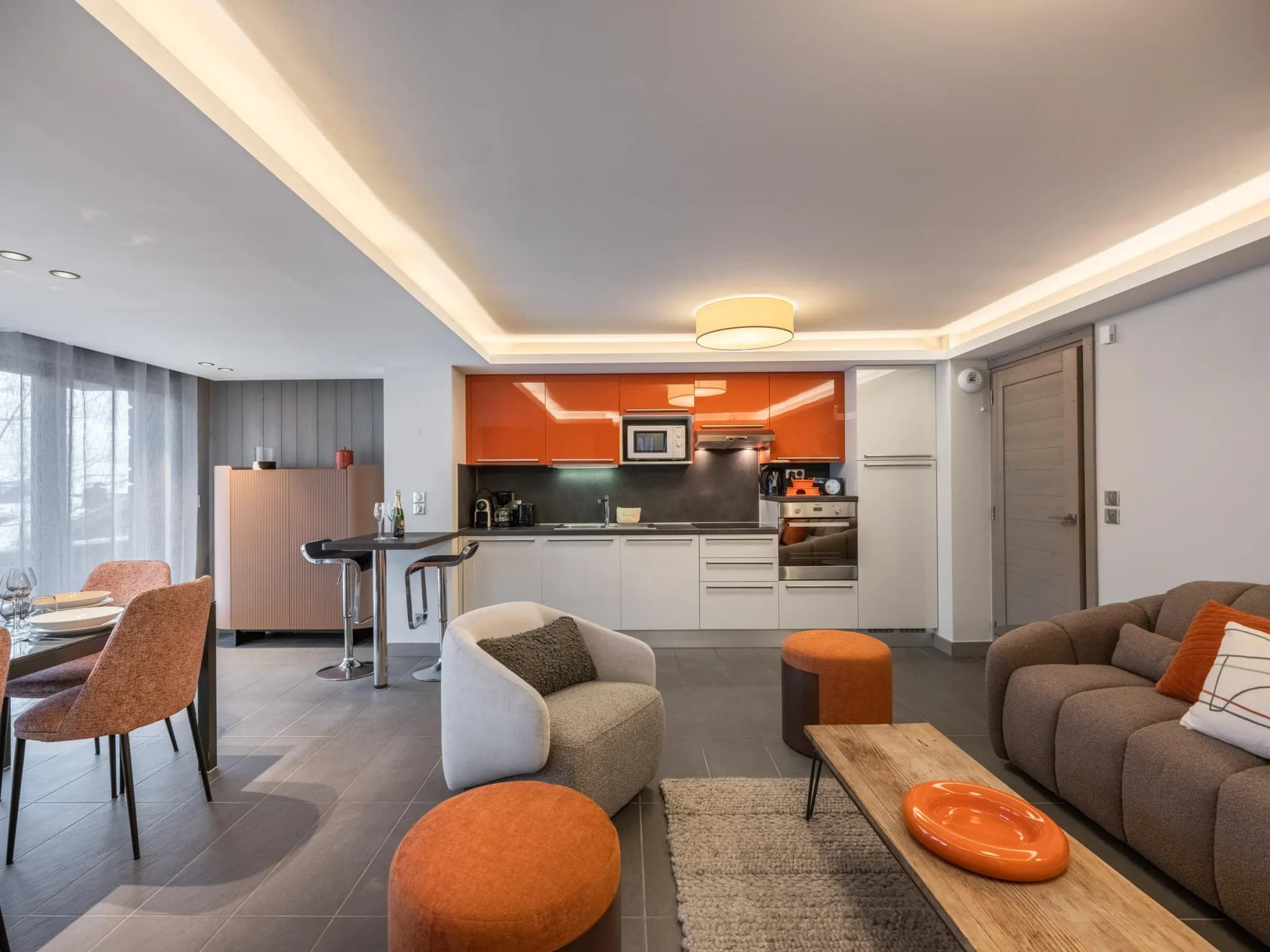 Open-plan living area with sofa, armchair, orange poufs and kitchen behind