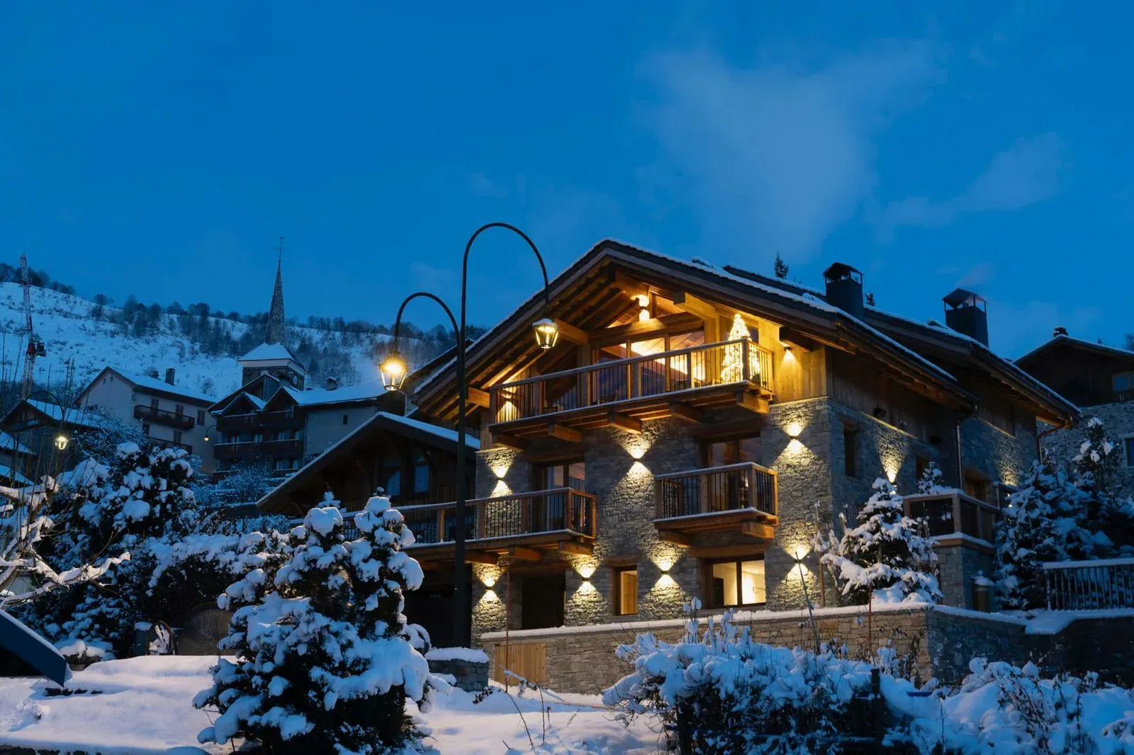 Stone and timber chalet at dusk with lit balconies and snowy village behind