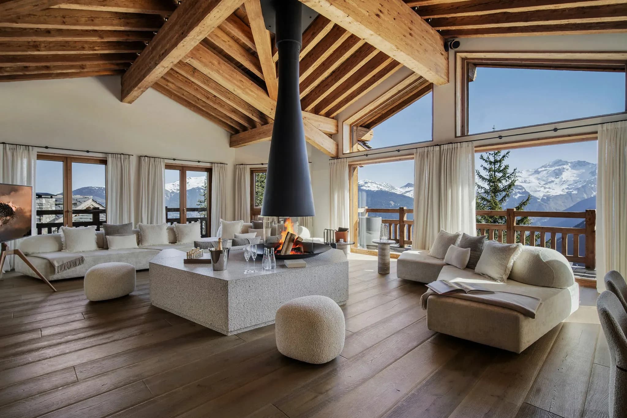Open-plan living room with central fireplace, exposed beam ceiling, and panoramic alpine views