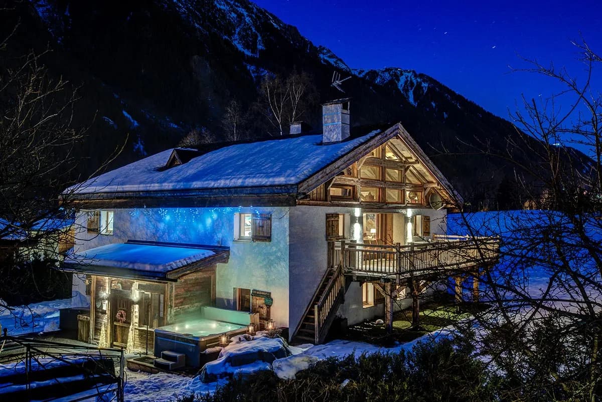 Snow-covered chalet illuminated at night, with outdoor hot tub and mountain backdrop