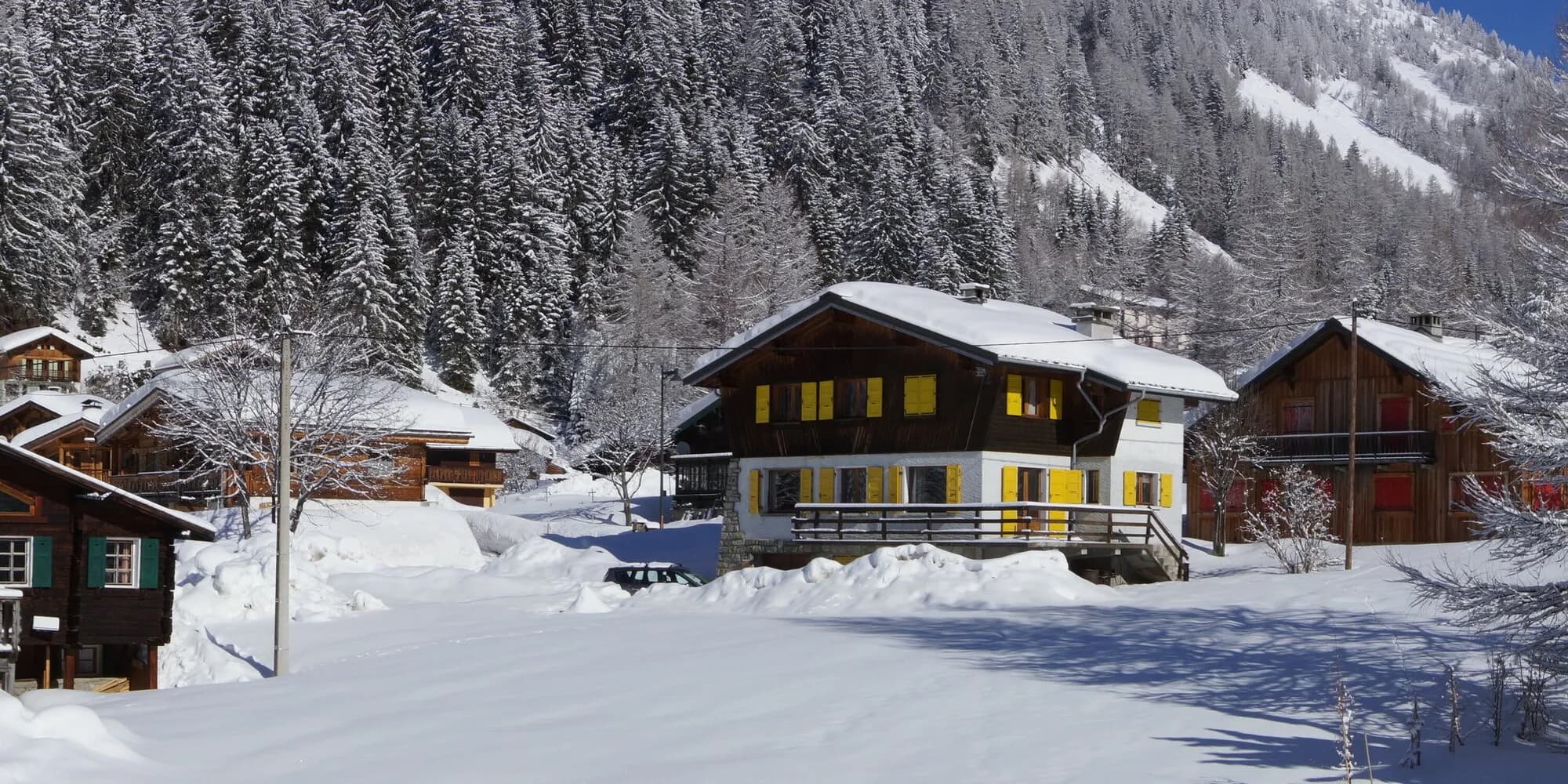 Wooden chalets with yellow and red shutters in deep fresh snow below a forested slope.