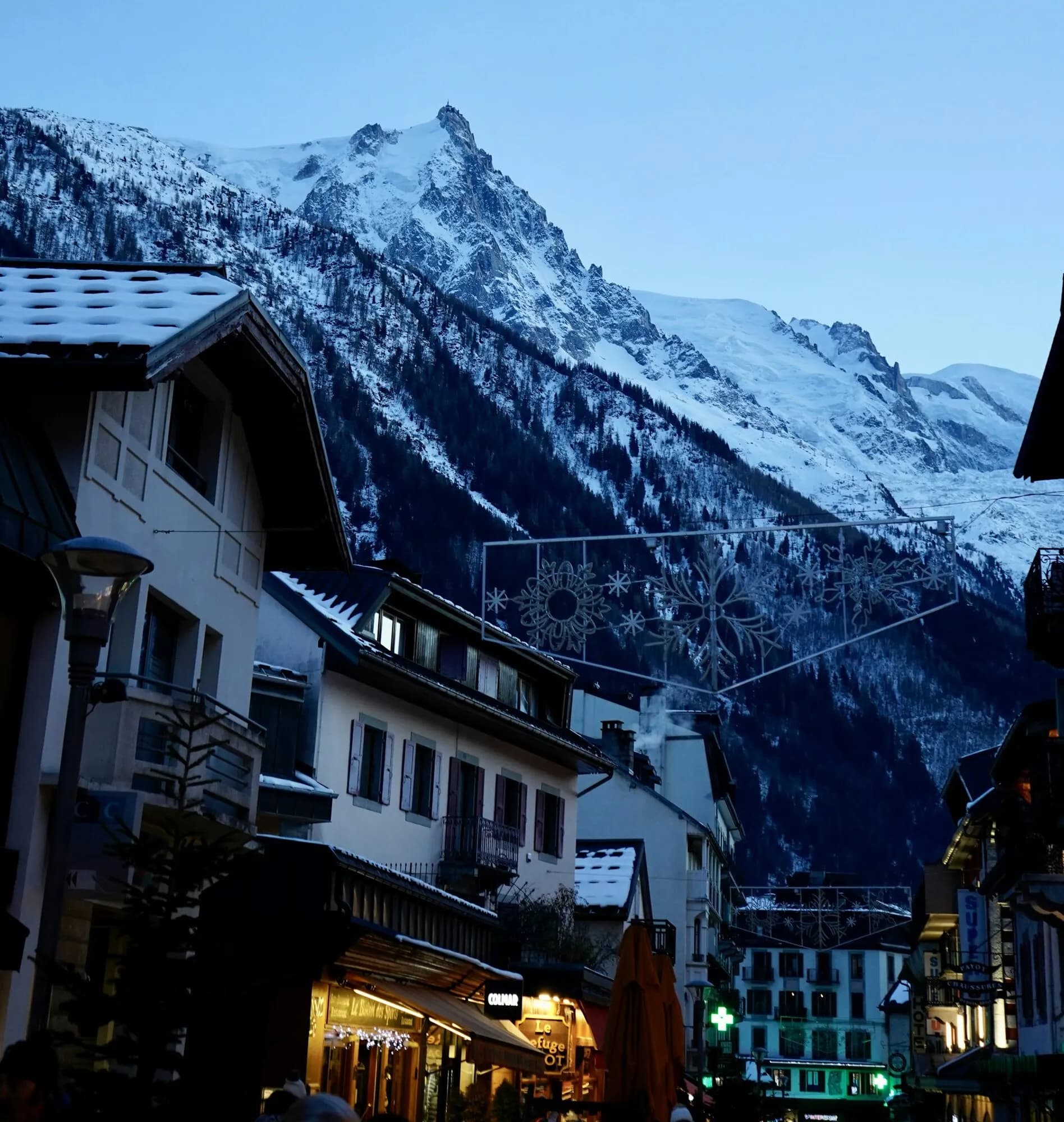 A snowy-roofed street in Chamonix at blue hour with a sharp granite peak above.