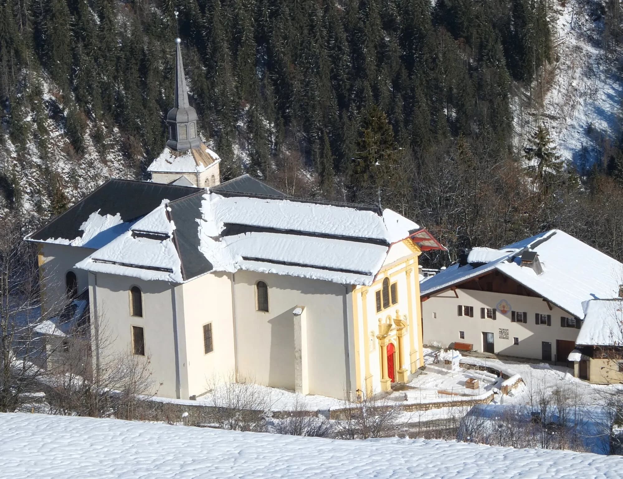 The baroque church of Saint-Nicolas-de-Véroce with snow on its roofs and the slope below, beside a chalet and fir forest.