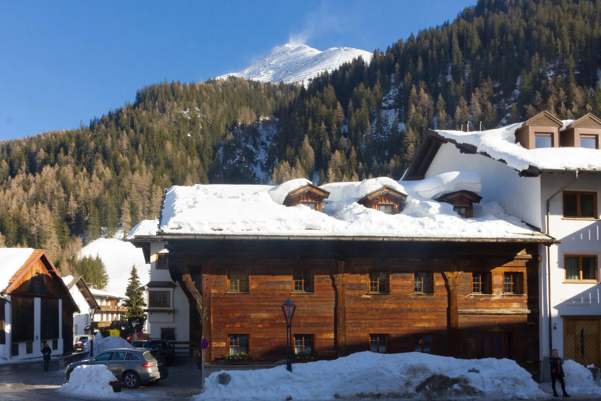A historic wooden house with a deep snow-laden roof below fir forest and a snowy summit.