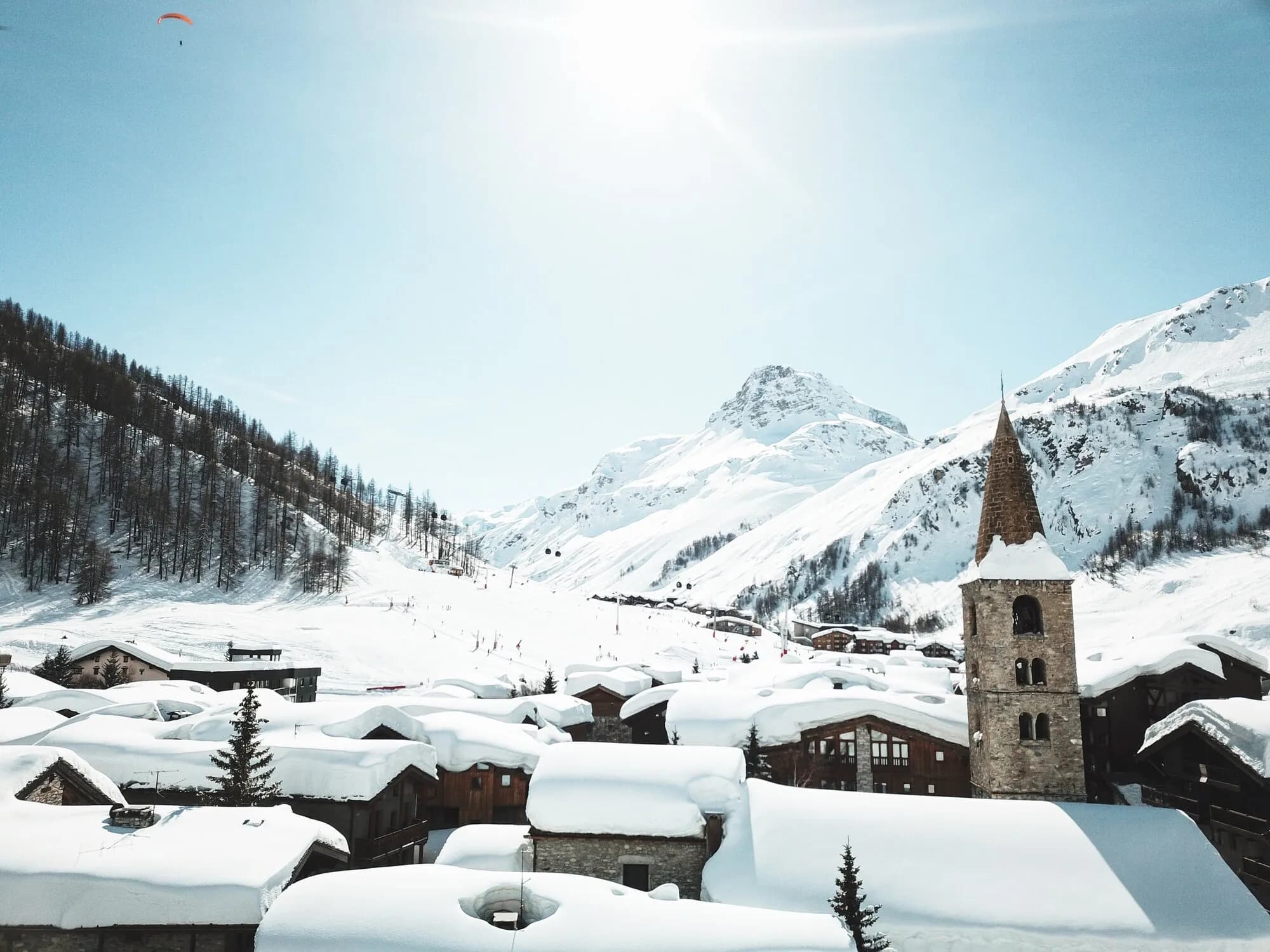 Deep snow on the roofs of the old village around the stone church tower, with snowy mountains behind.