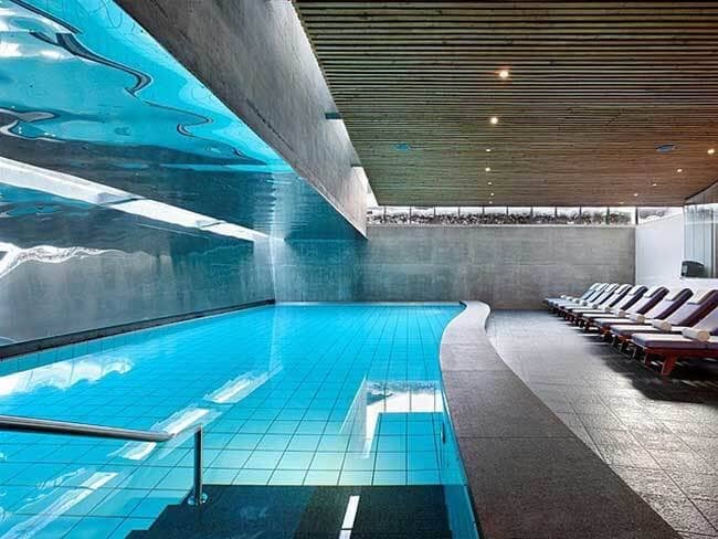 Indoor heated pool with poolside lounge seating and wood-slat ceiling