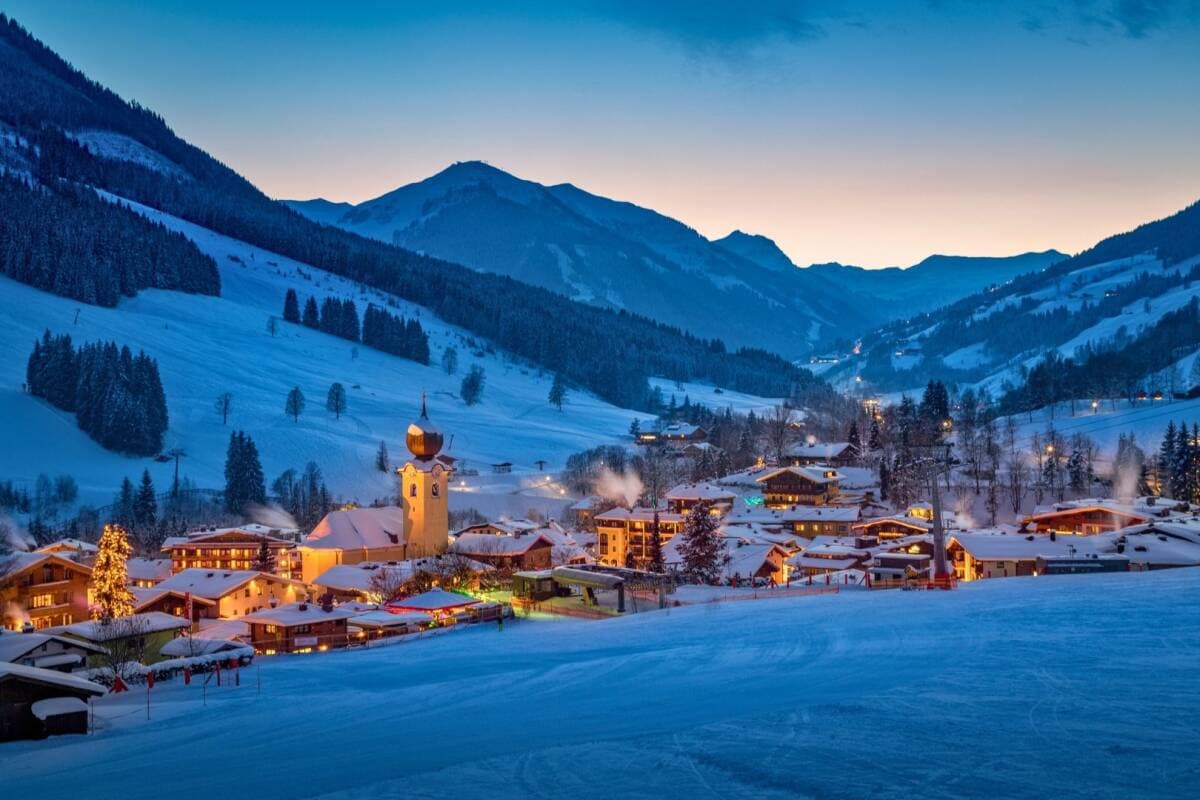 Alpine village at dusk with church tower, frozen lake, and ski-slope mountain backdrop